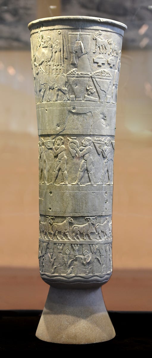 Close-up of the tall, thin Warka Vase or Uruk Vase, glowing rich amber under museum lights. Vertical sides with a slight opening toward the top, covered by carved images. Close-up of the tall, thin Warka Vase or Uruk Vase, glowing rich amber under museum lights. Vertical sides with a slight opening toward the top, covered by carved images.