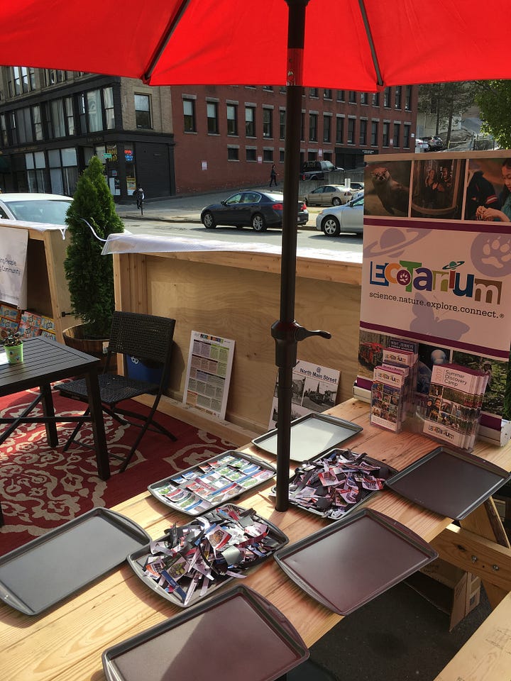 Left image: A photo of the Magnetic Neighborhood interactive: A cookie baking tray covered in magnetic pictures of city elements like streets, parks, and different types of buildings. The baking sheet is being put into a scanning device that will record a picture of the neighborhood on the tray. Right image: The Magnetic Neighborhood magnets and cookie trays, set on a picnic table in a wood-enclosed “parklet” on a city street.