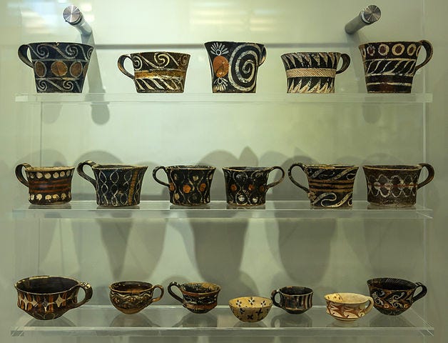 Three clear plexiglass museum shelves filled with ancient Minoan loop-handled cups in different sizes and shapes, most of them painted in geometric designs in the colors black, white, and red.