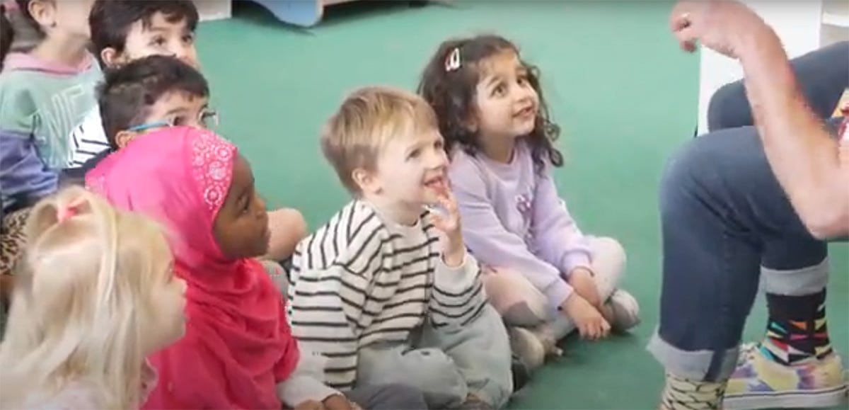 Children aged 5 and 6 with expressions of gleeful engagement, sat on a carpet in front of Storyteller Chip