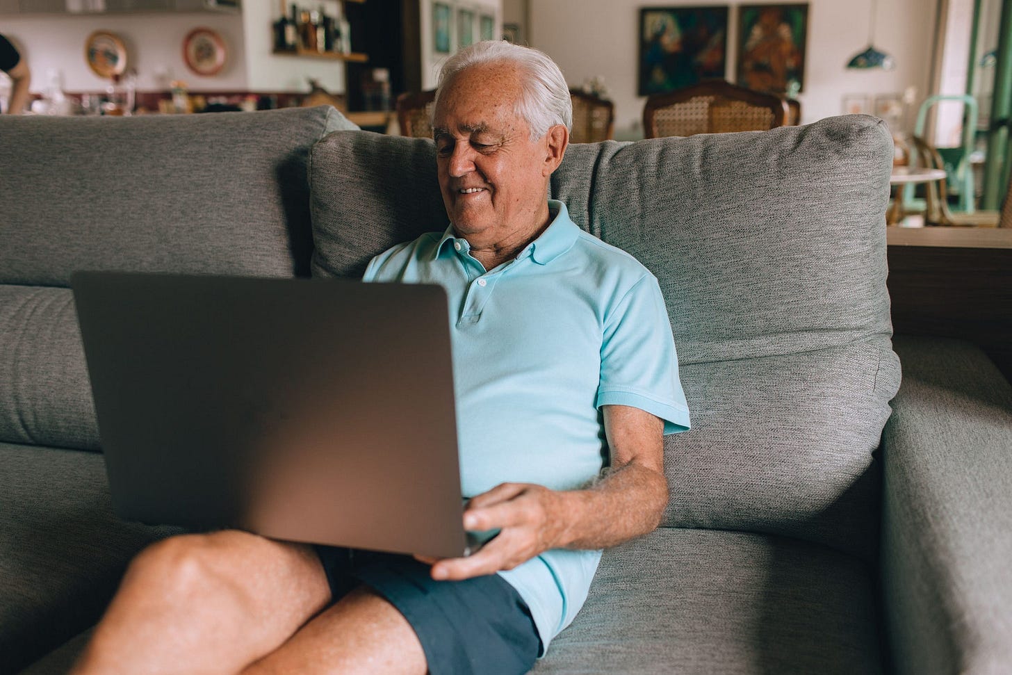 Senior man in blue polo sitting on couch using laptop in a cozy living room setting.