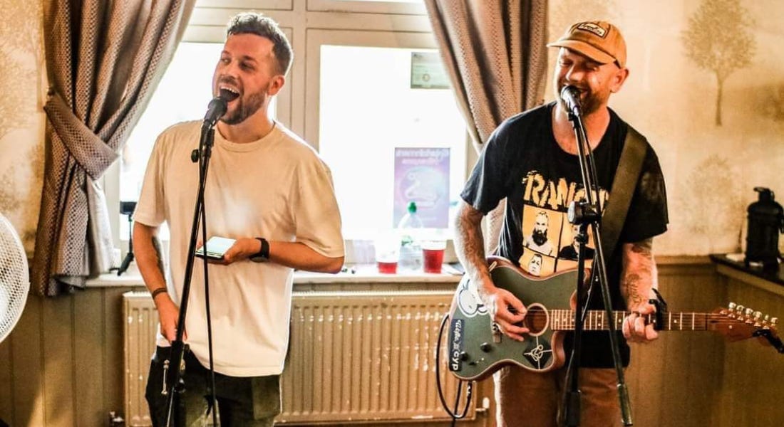 Two men singing in front of microphones with beige walls, brown curtains around a window, and a white radiator behind them Two men singing in front of microphones with beige walls, brown curtains around a window, and a white radiator behind them