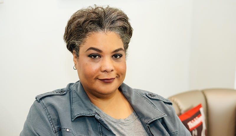A Black woman wearing a jean button-up shirt and grey undershirt, smiles coyly into the camera as she looks directly into your soul like she knows all your secrets.