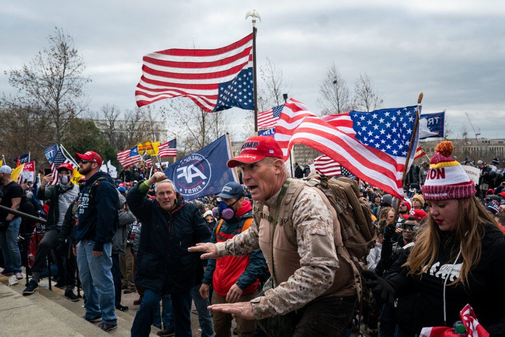 Ray Epps, in the red Trump hat, center, gestures to a line of law enforcement officers, as people gather on the West Front...