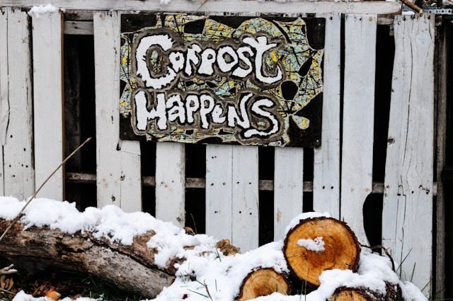 photograph of white picket fence with a handprinted sign that reads "Compost Happens" and a woodpile with snow
