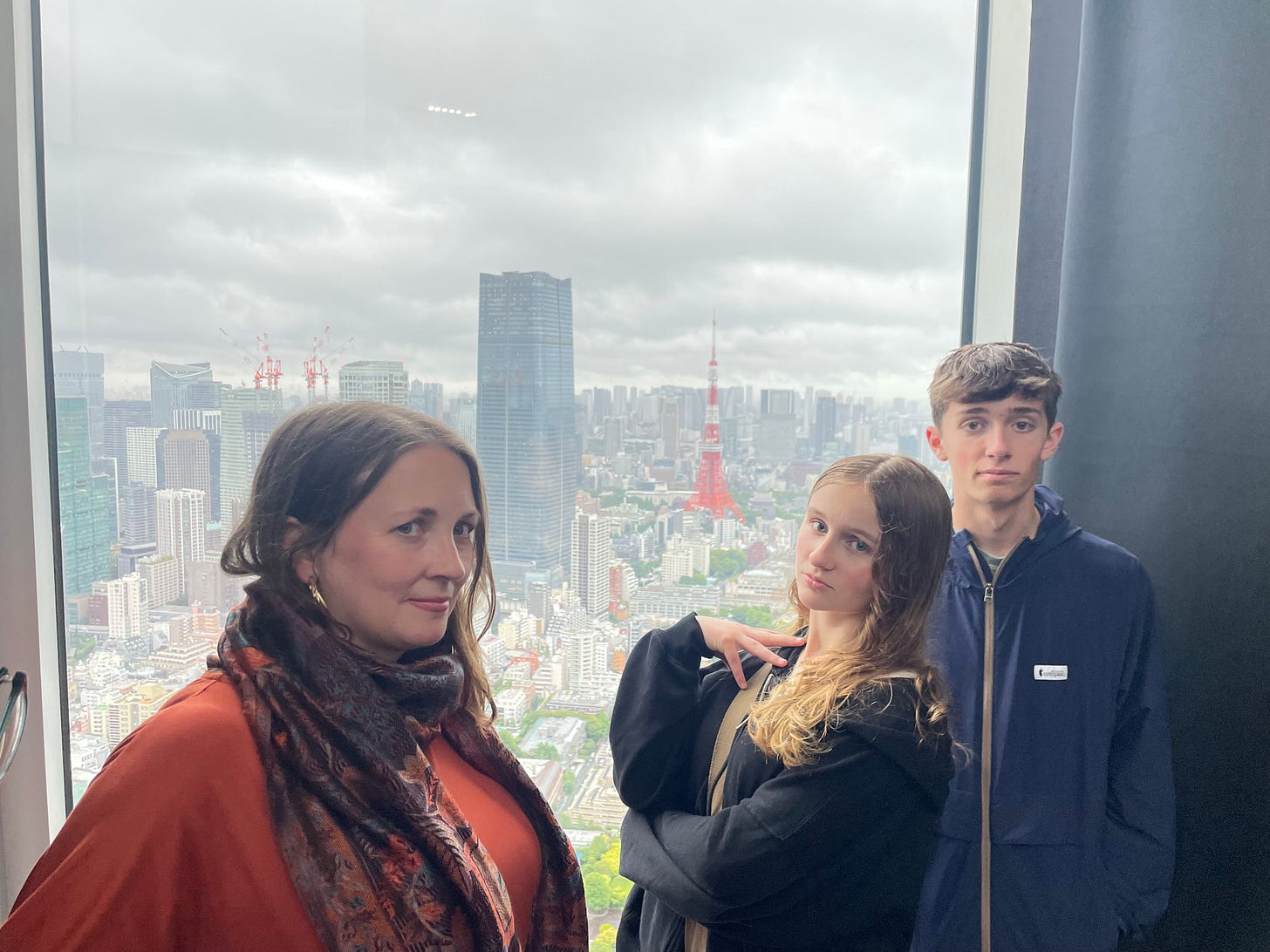 A middle aged woman in an orange shirt, a teenaged girl in a black jacket, and a teenaged boy in a blue jacket stand in front of a window overlooking the Tokyo skyline. It is cloudy.