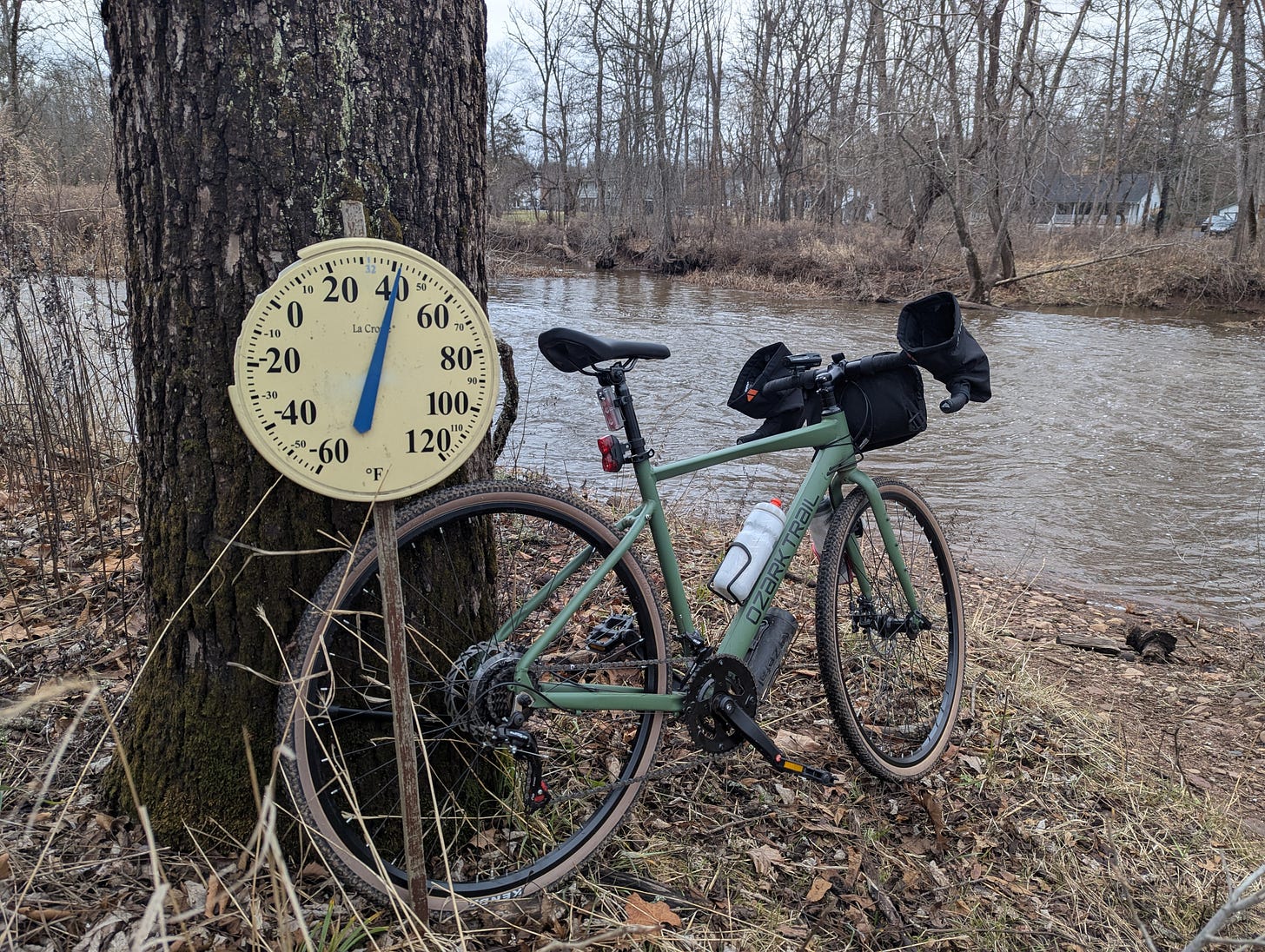 Winter photo alongside a small river. A thick tree stands on the riverbank. A large thermometer is attached to the tree. The thermometer indicates that it is 40 degrees F. Leaning on the tree is a green bicycle. It is pointing towards the river. On the opposite bank are leafless trees. The photo is mostly browns.