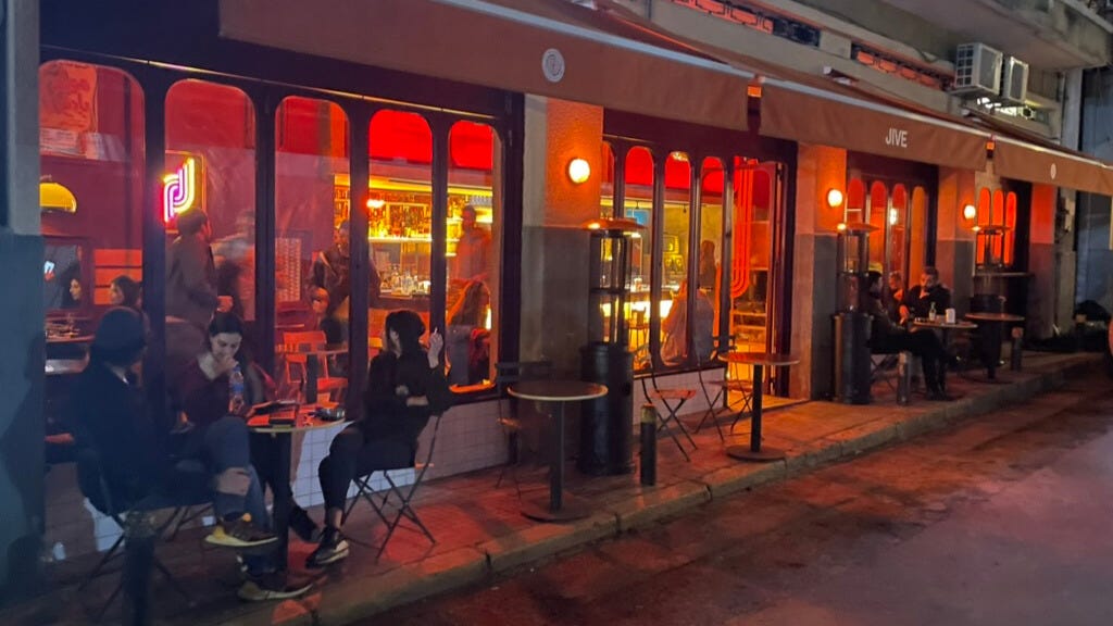 The outside of a brightly lit bar. People sit out front smoking cigarettes. 