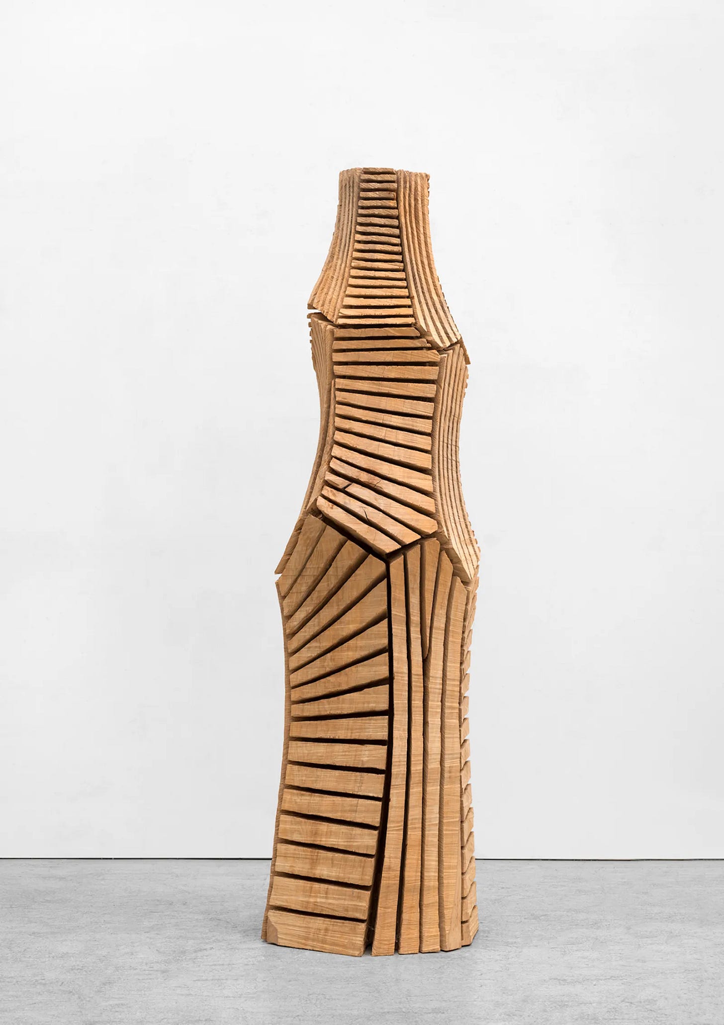 Tall geometric wooden sculpture by David Nash with stacked carved segments and rhythmic linear cuts, photographed against a pale gallery wall. Tall geometric wooden sculpture by David Nash with stacked carved segments and rhythmic linear cuts, photographed against a pale gallery wall.