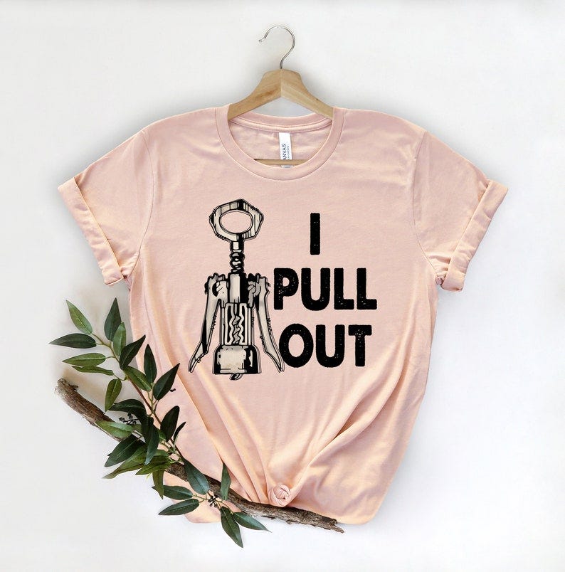 May include: Peach-colored t-shirt featuring a graphic of a corkscrew and the text 'I PULL OUT' in black. The shirt is displayed on a hanger against a white background.  The design is simple and features a vintage-style corkscrew illustration. The shirt appears to be made of soft, lightweight material, suitable for casual wear.  It's a fun, wine-themed tee perfect for wine lovers.