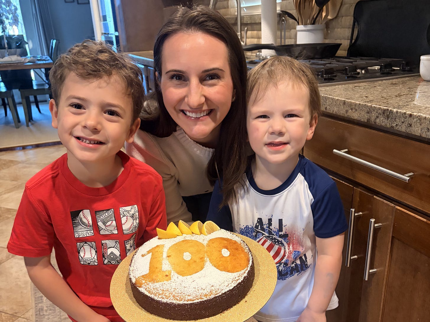 A mom and her two young boys smiling while holding a cake with the number 100 on it
