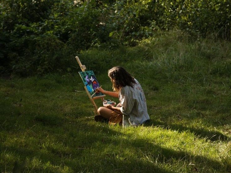 This may contain: a woman sitting in the grass painting on an easel