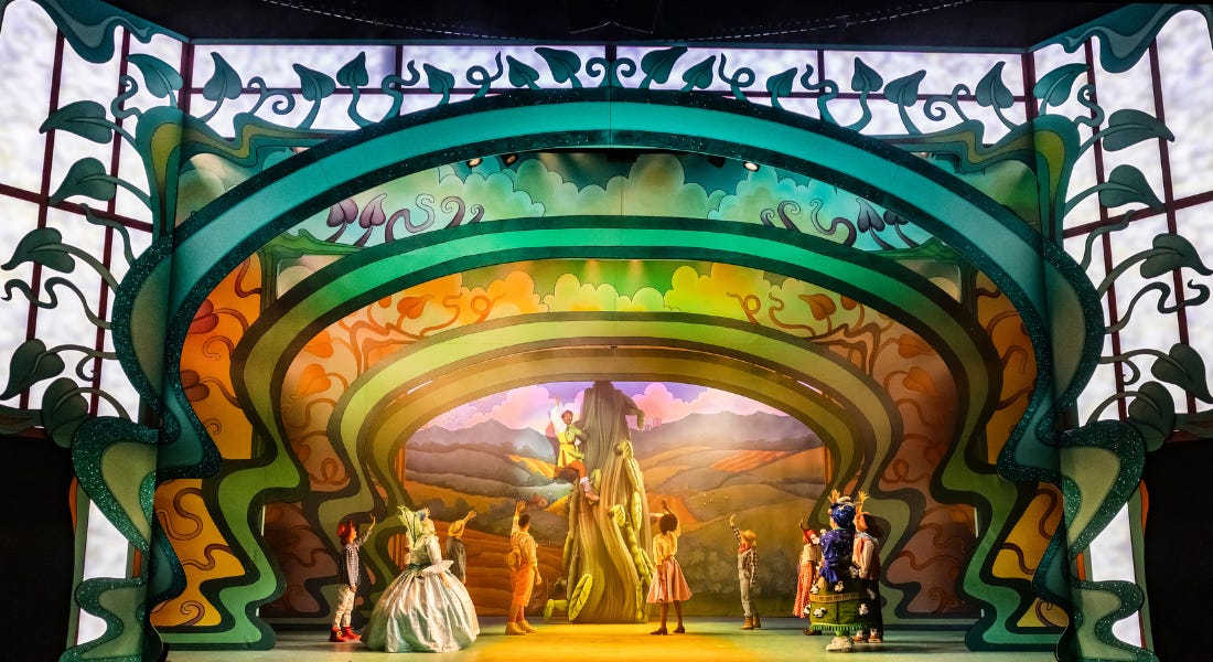 A wide shot of a stage, filled with shades of green and orange. On the stage, nine performers, in a variety of colourful costumes, stand waving. Towards the back of the stage, a man is climbing a giant inflatable beanstalk. He is waving to those on the ground. 
