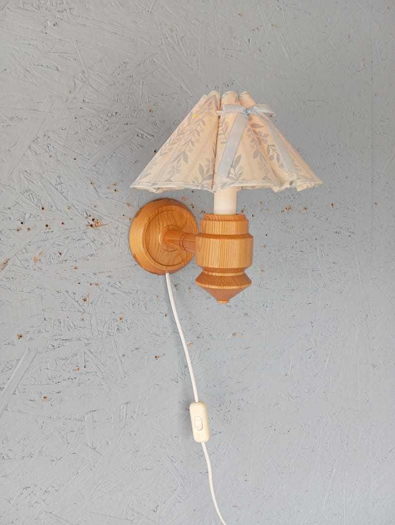 May include: A wooden wall lamp with a light blue and white floral fabric lampshade. The lamp has a white cord with an inline switch.