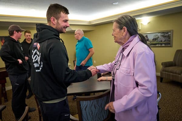 A young man in a black hooded sweatshirt smiles while he shakes hands with an older woman in a purple jacket. 