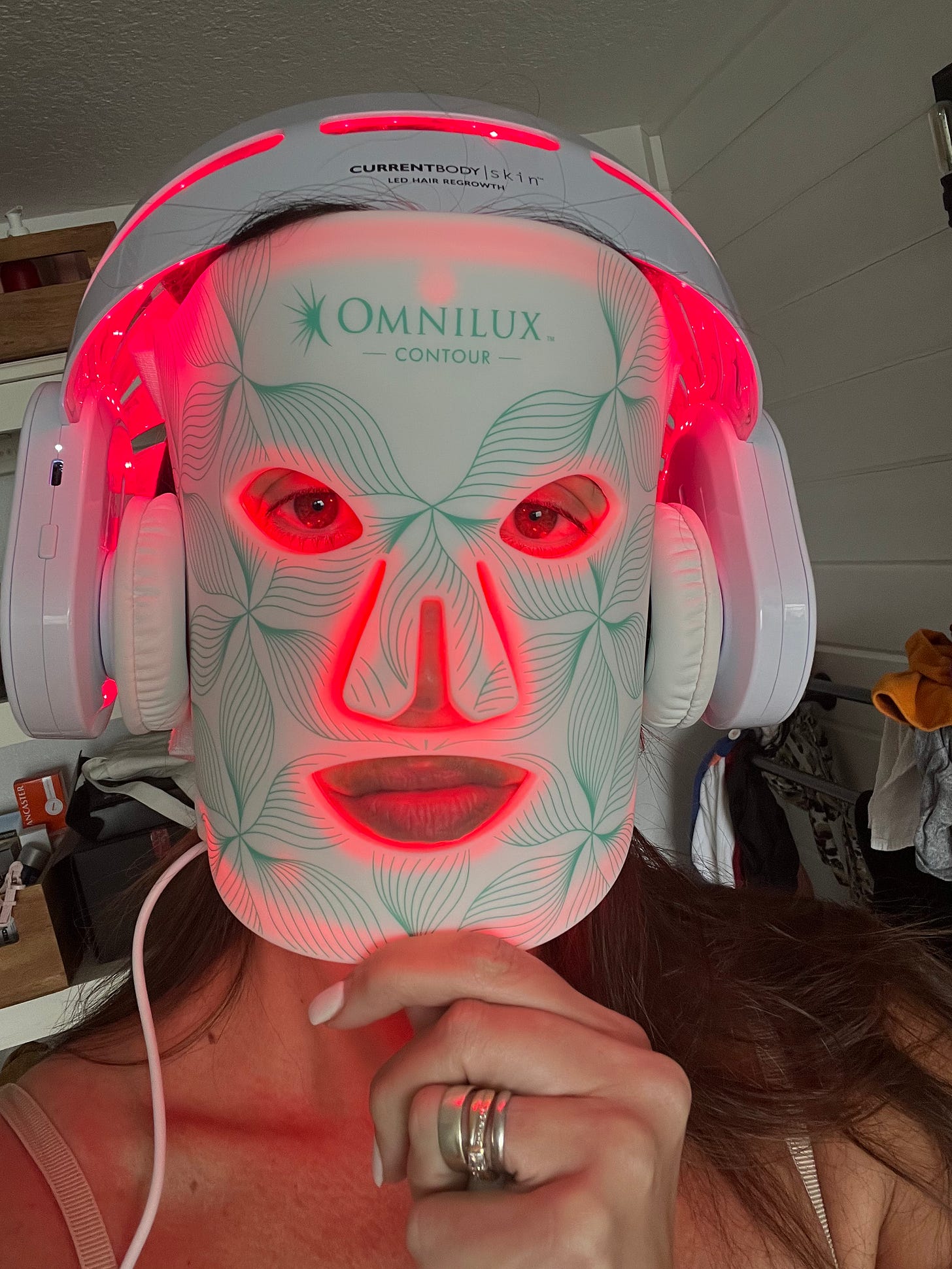 Close-up of a woman wearing a glowing Omnilux LED face mask underneath a Current Body red light hair growth helmet. Close-up of a woman wearing a glowing Omnilux LED face mask underneath a Current Body red light hair growth helmet.