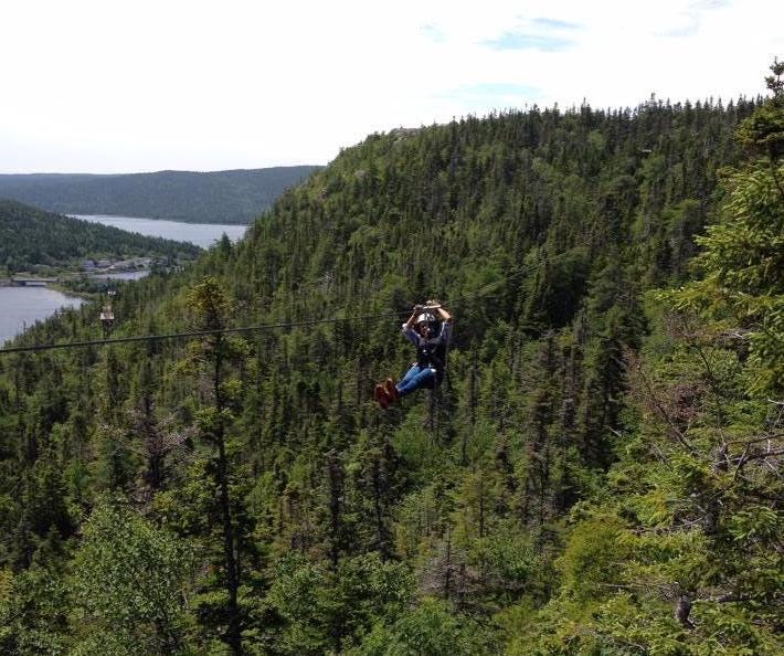 A small, helmeted figure zooms across a forested valley on a zip line, against a background of sea and sky.