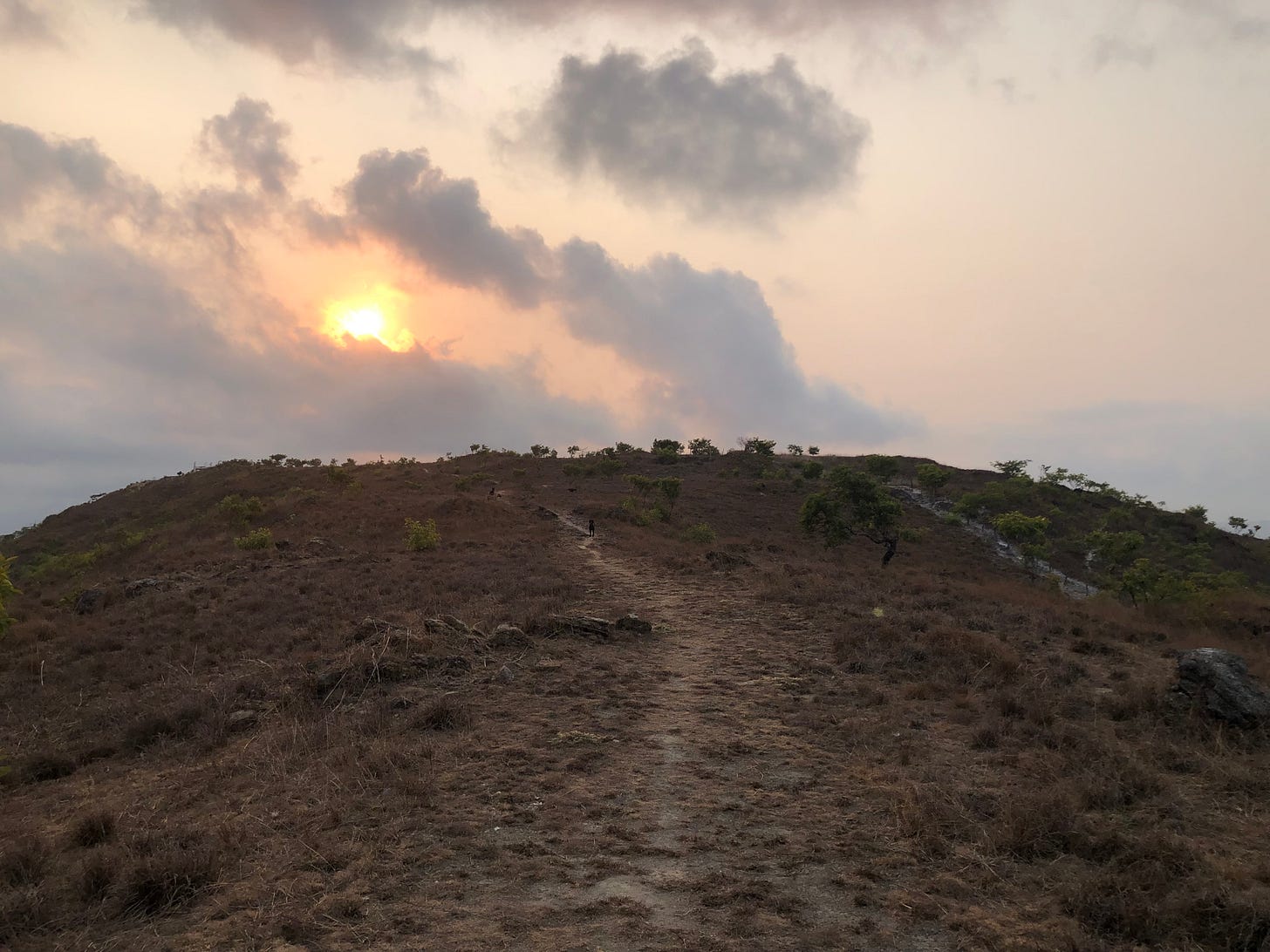 A hill covered in dead grass with a faded path down the middle, a sunset and runny clouds in the background.