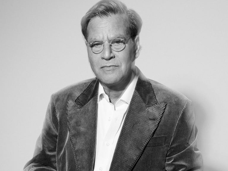 A man with glasses and a striped jacket stands with hands clasped in front of a light gray background.