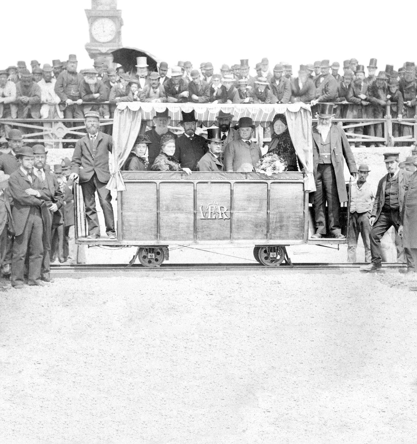 Monochrome photographic print showing the opening of Volk's Electric Railway. Shows numerous dignitaries inside car. Large crowds in attendance, both to sides of car and on promenade above. Clock tower of Aquarium visible in background.