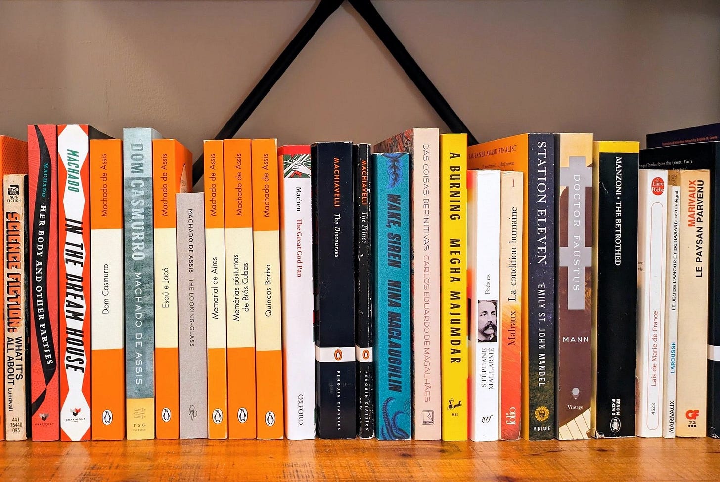 Photo of a section of a bookshelf with various bookspines visible, some in Portuguese, some in French, the majority in English. There's a concentration of bright orange spines at left, all by Machado de Assis. Photo of a section of a bookshelf with various bookspines visible, some in Portuguese, some in French, the majority in English. There's a concentration of bright orange spines at left, all by Machado de Assis.
