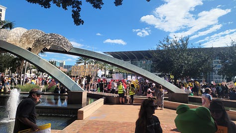 20251018_114518.jpg: A close-up of a hand-drawn protest sign reading "NO KINGS!!" and "NO WEENIES ALLOWED," featuring a crossed-out Burger King logo and a drawing of SpongeBob SquarePants.  20251018_113815.jpg: A wide shot of a large, sunny protest in a public plaza, showing crowds gathered around a modern arched sculpture and a water fountain.  20251018_113805.jpg: A crowd of protestors gathered on a brick plaza under a canopy of trees, with some people holding American flags and various signs.  20251018_110151 - Copy.jpg: A person in an inflatable blue pterodactyl costume stands at the protest, next to a person in a wheelchair holding a sign that reads, "DECLARE INDEPENDENT. FLUSH THE RED/BLUE WAR!"  20251018_113823.jpg: A wide view of the protest centered on a large, modern arched sculpture over a fountain, with a large crowd and many signs visible in the background.