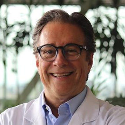 Homem de meia-idade, de cabelos escuros e grisalhos, usando óculos de armação preta e jaleco branco sobre uma camisa azul. Ele aparece sorrindo para a câmera. Homem de meia-idade, de cabelos escuros e grisalhos, usando óculos de armação preta e jaleco branco sobre uma camisa azul. Ele aparece sorrindo para a câmera.