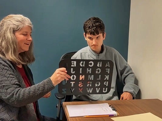 A practitioner holds a black letterboard stencil as a young man seated at a desk looks toward it A practitioner holds a black letterboard stencil as a young man seated at a desk looks toward it