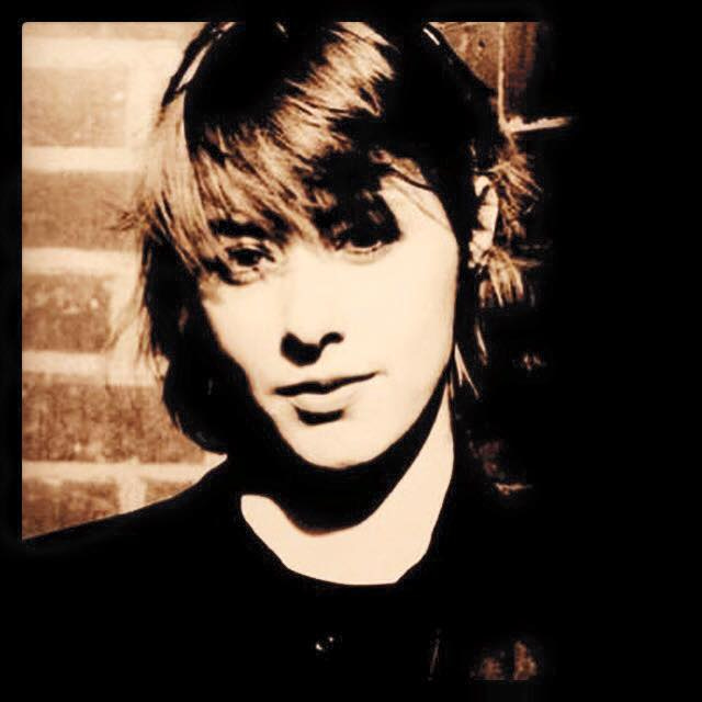 Suzanne Vega, PR shot 1985, left, with assymetrical new-wave haircut against brick wall; at Lula Lounge on March 11, right, singing into microphone with long hair in top hat and black tuxedo-pantsuit, with sparkly dangly chain-link necklace. Her acoustic guitar is on a stand beside her.