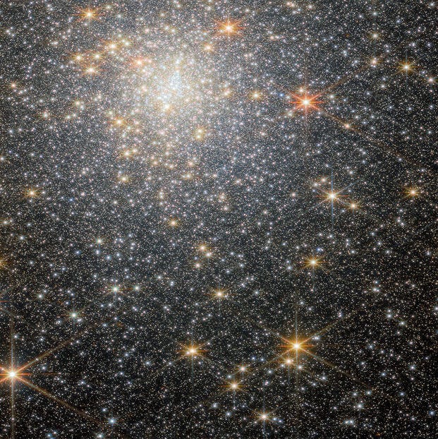 A dense star field with numerous stars of varying brightness. The stars range from white to yellow and orange, creating a glittering, speckled effect against the dark background of space. The brightest stars are located towards the top left, where they form a loosely concentrated cluster.