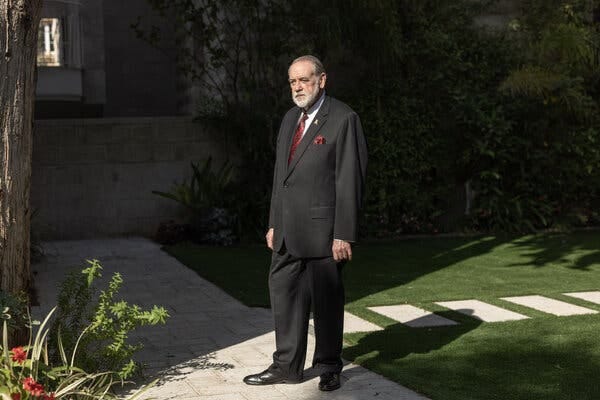 A man in a dark suit standing on a walkway with grass and shrubbery in the background. A man in a dark suit standing on a walkway with grass and shrubbery in the background.