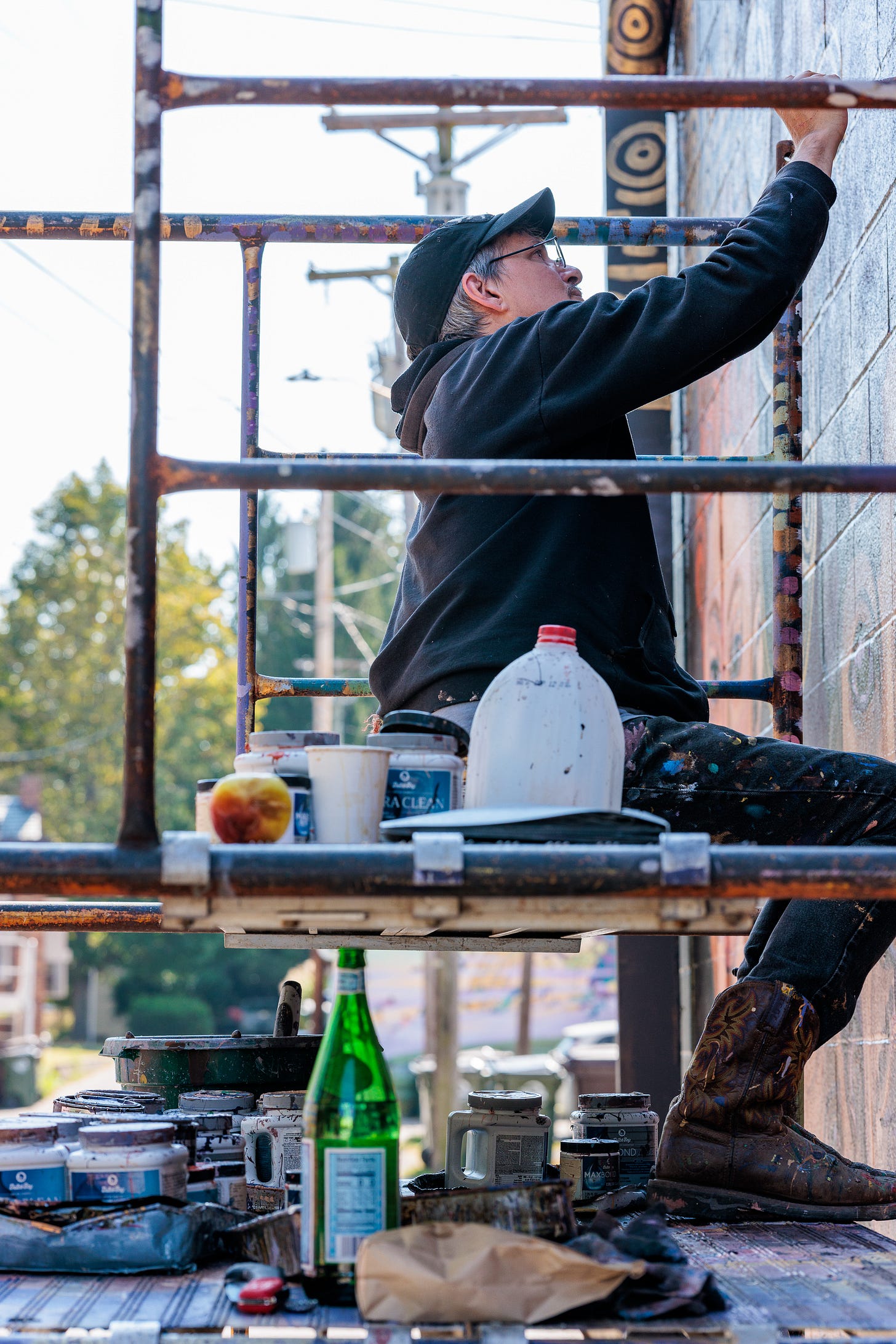 Pierre Nagley sits on a scaffold plank reaching up to paint, surrounded by paint jars, a water bottle, and a rag on the platform below him.