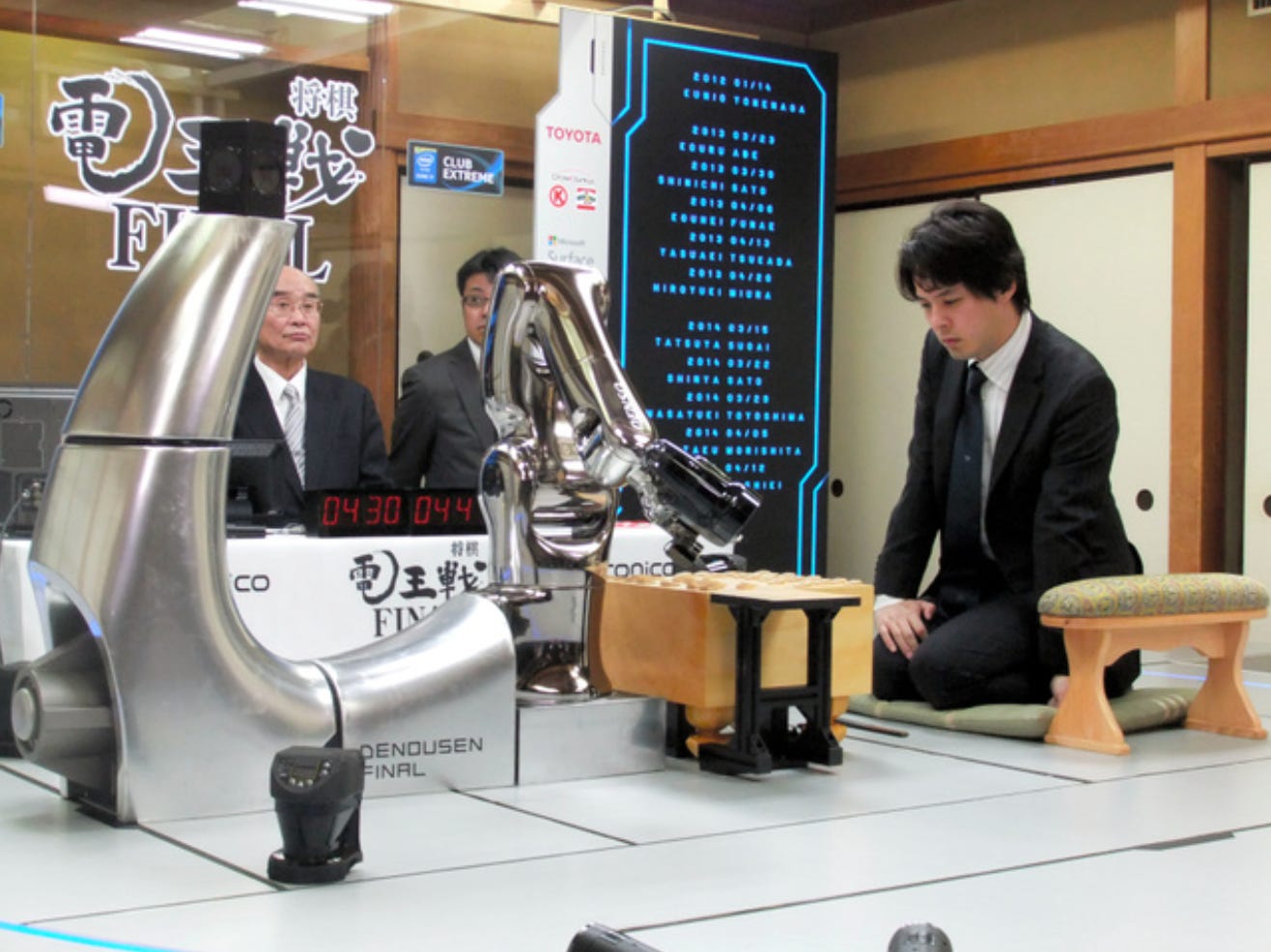 A scene from Denō-sen. In the middle of the frame is a shogi board. To the right, a man in a suit sits on a cushion. To the left, there is a robotic arm extending toward the board. A scene from Denō-sen. In the middle of the frame is a shogi board. To the right, a man in a suit sits on a cushion. To the left, there is a robotic arm extending toward the board.
