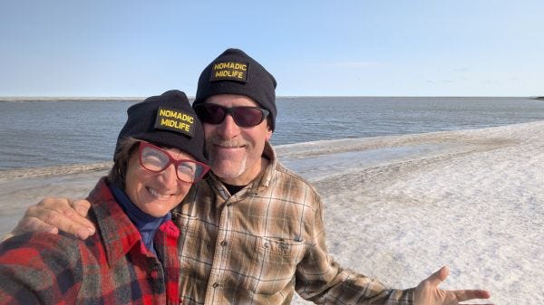 two smiling people standing on a dirty bank of old snow in front of a large body of water
