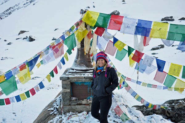 White woman with blond hair is wearing colorful yak-knit hat and is standing in front of a memorial with prayer flags. She is surrounded by snow. 