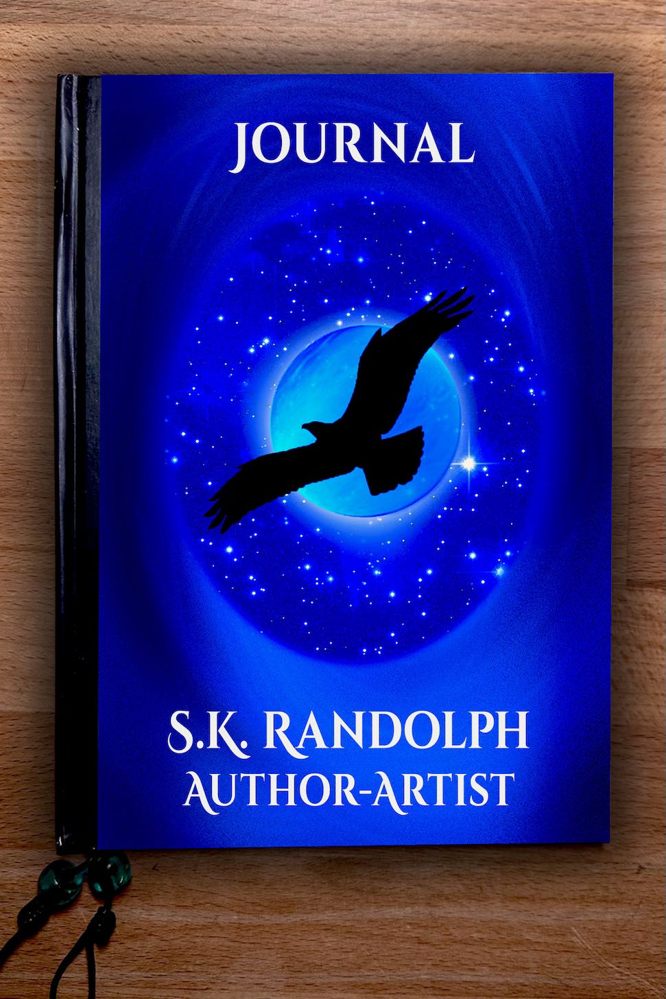 A journal with a blue cover featuring a silhouette of a flying bird against a starry sky and glowing moon. The text reads JOURNAL at the top and S.K. RANDOLPH AUTHOR-ARTIST at the bottom.