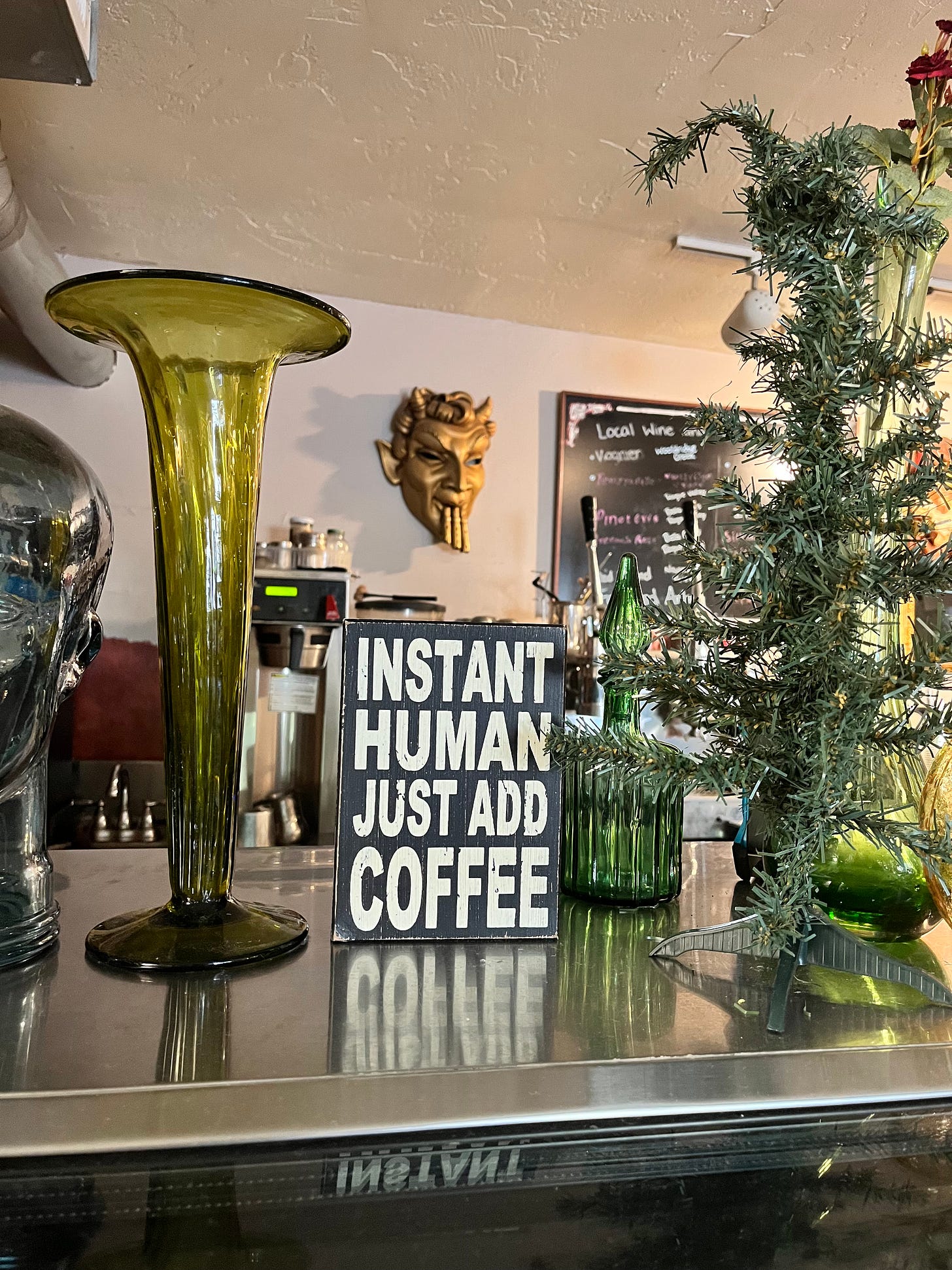 a bar at a coffee house with a glass vase and sign that says Instant Human Just Add Coffee and above it is a mask, probably Pan with his flute