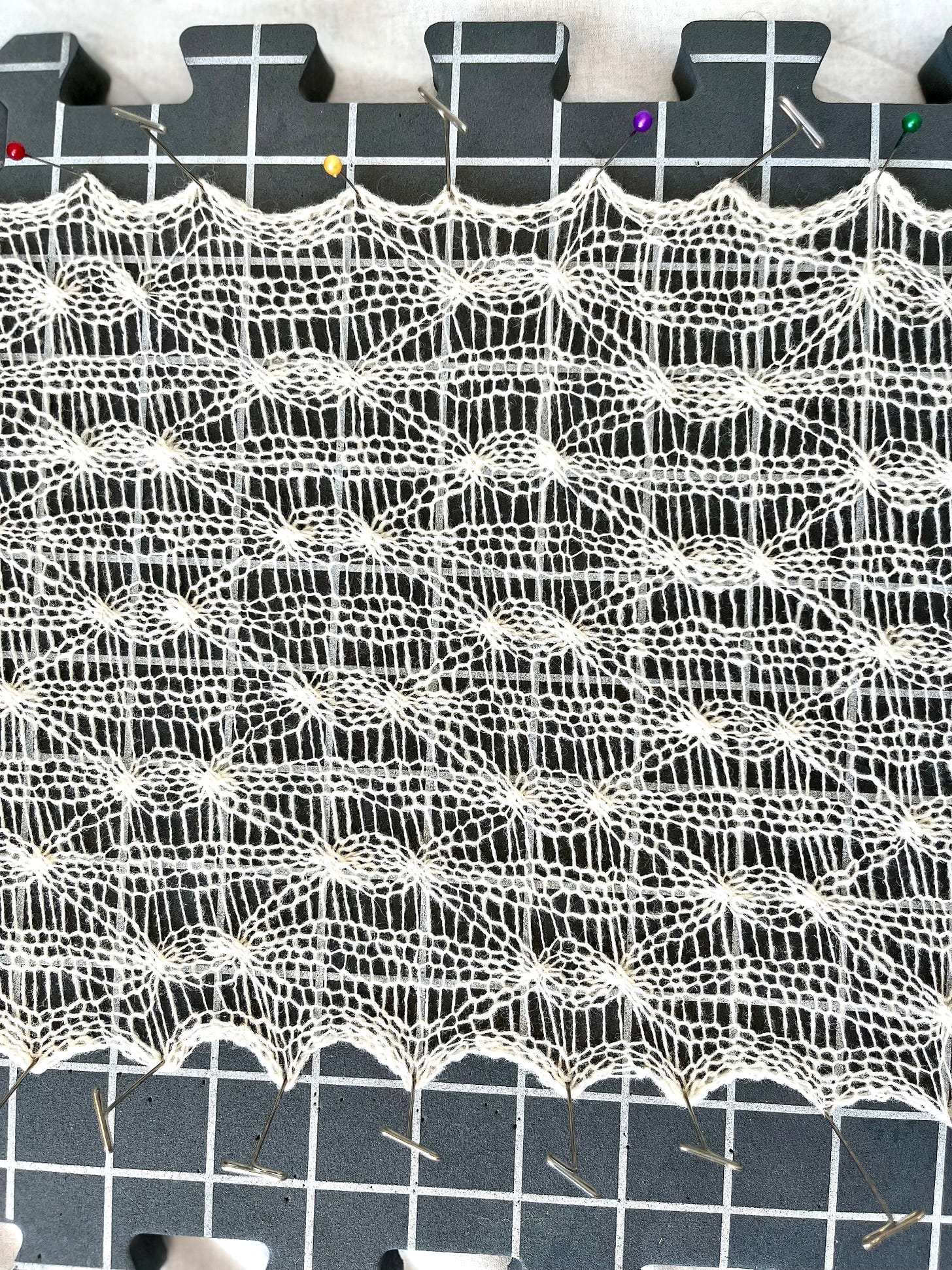 Close-up of a cream lace knitted fabric pinned to a grey foam blocking mat. The pattern shows elongated diamond shapes formed by 2x2 cables and vertical ladder openings running the length of the fabric.