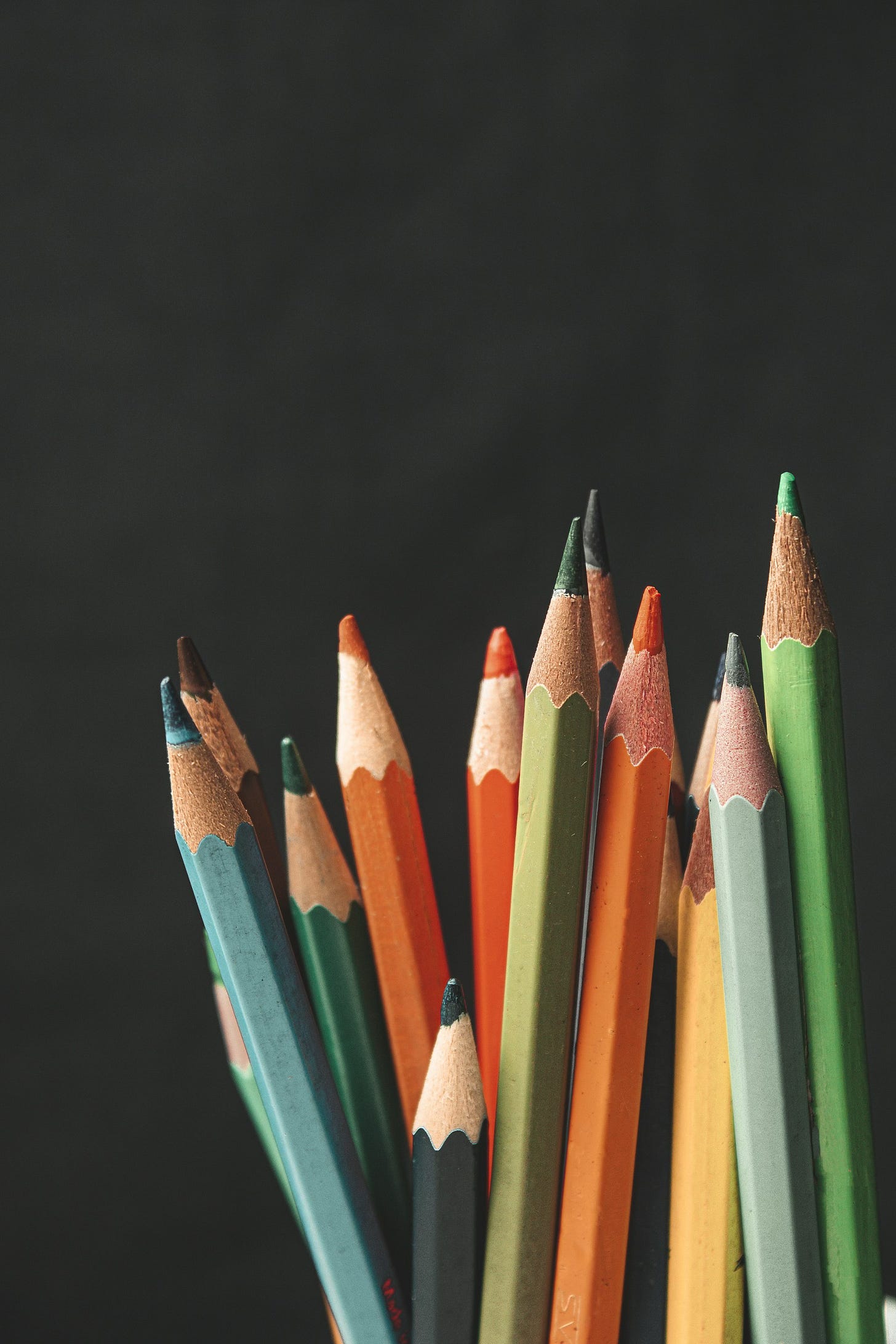 A handful of sharpened colored pencils, mostly in greens and oranges, stand upright against a black background.