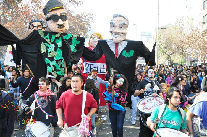A slightly overexposed, washed-out digital image of a student protest marching down a wide street, probably in downtown Santiago. In the foreground, young people walk playing large drums and cymbals, a sort of popular marching band known as a "batucada." Just behind them loom four giant papier mache puppets—each two people tall—carried by other students on poles. The two in front are former dictator Augusto Pinochet on the left, and president Sebastián Piñera on the right. They are dressed in all black and grin evilly, pockets full of money. Behind them are two other puppets, partially obscured, and students marching out as far as can be seen. A slightly overexposed, washed-out digital image of a student protest marching down a wide street, probably in downtown Santiago. In the foreground, young people walk playing large drums and cymbals, a sort of popular marching band known as a "batucada." Just behind them loom four giant papier mache puppets—each two people tall—carried by other students on poles. The two in front are former dictator Augusto Pinochet on the left, and president Sebastián Piñera on the right. They are dressed in all black and grin evilly, pockets full of money. Behind them are two other puppets, partially obscured, and students marching out as far as can be seen.