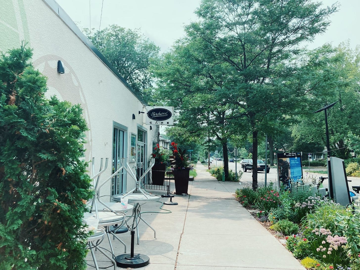 the front of birchwood cafe, a white building with a white and black sign reading "Birchwood Cafe", surrounded by lush green trees and bushes, and metal chairs and tables stacked to the side