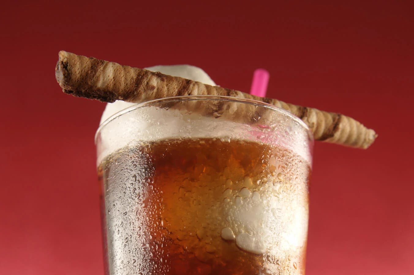 A imagem exibe o closeup de um copo de refrigerante de root beer contra um fundo vermelho. A bebida é decorada com um tubo de biscoito de canela e um canudo rosa-choque.