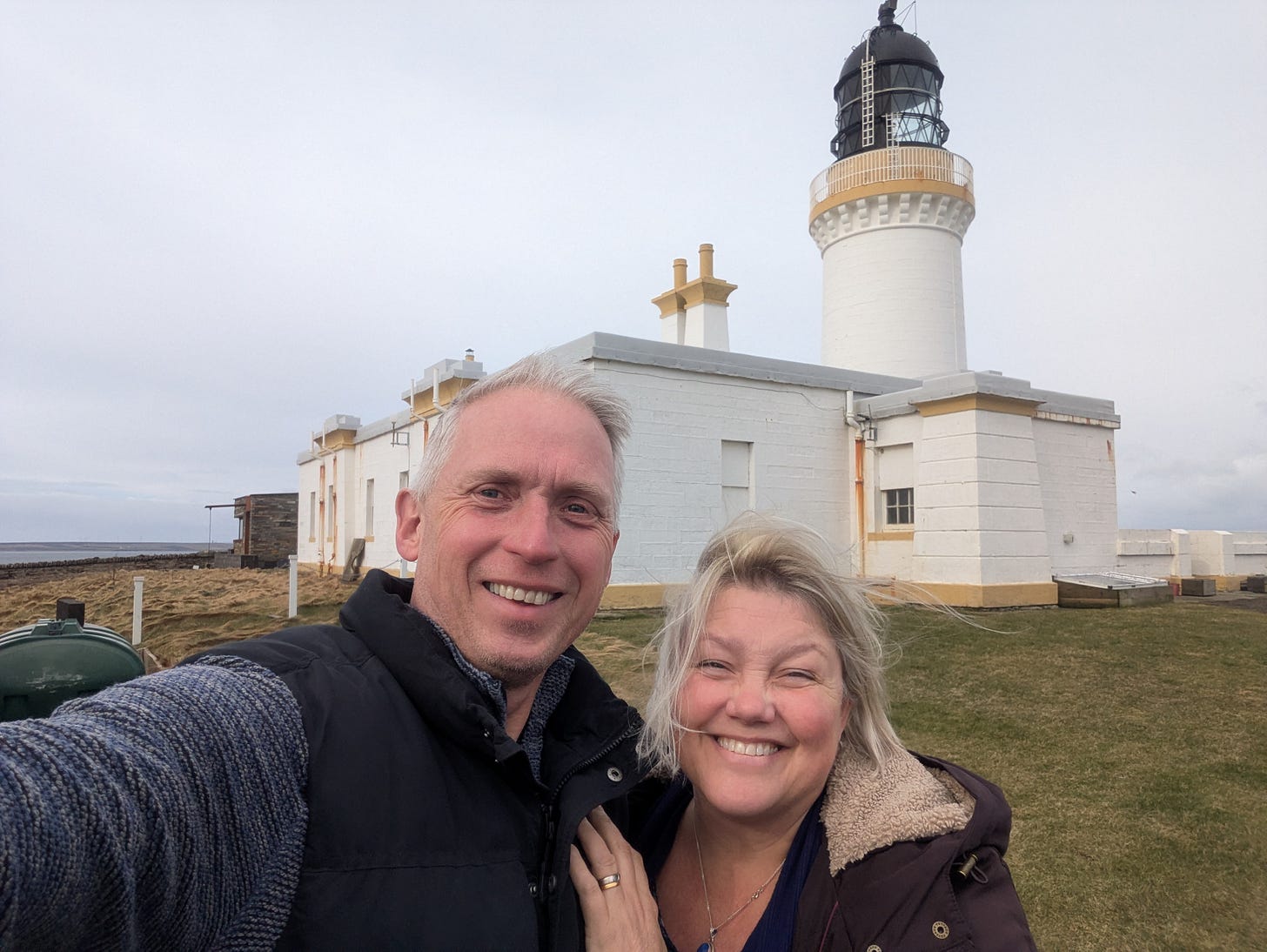 A selfie of Neil and Lucy smiling happily in front of the tall, white Noss Head Lighthouse and its attached keeper's cottage. The sky is slightly overcast and the coastal wind is blowing Lucy's hair.