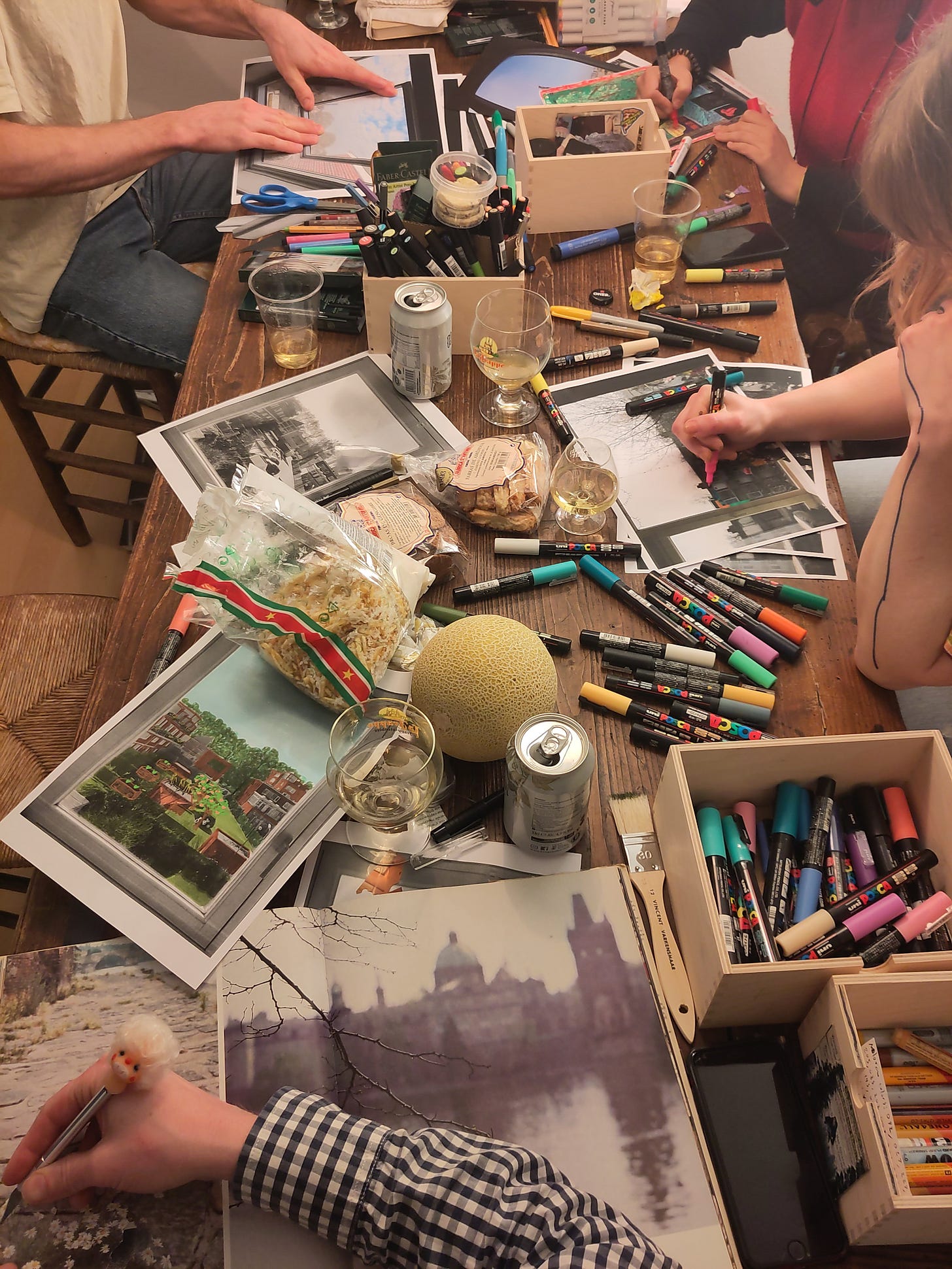 A dining room table covered in paper, pens, drinks, and food, messily arranged. Four sets of hands appear around the table, in the process of making art.