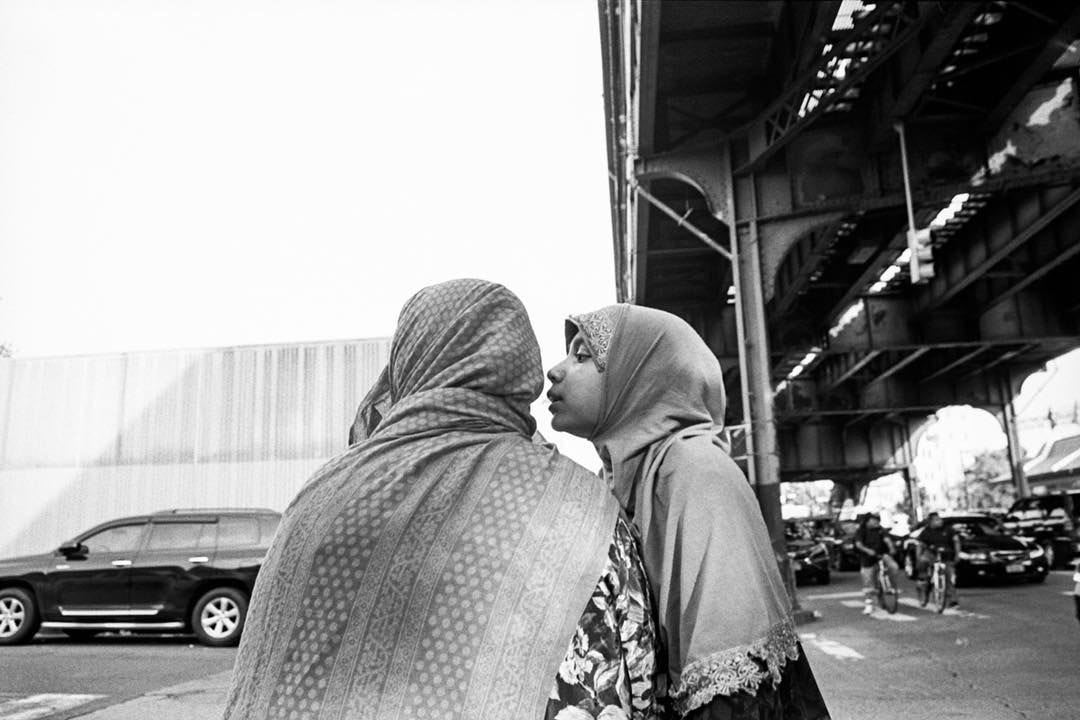 Black and white photo of a young girl wearing a hijab leaning over to another figure who is also wearing a hijab. We only see her back, so we have no idea how old this second figure is, or her relation to the girl. We can speculate that she must be a close relative because of the intimacy and closeness of the girl and the words we can only guess being whispered. In the background, there is a cast iron elevated railway (which we can only see the underside) making it likely that this is somewhere in New York City.