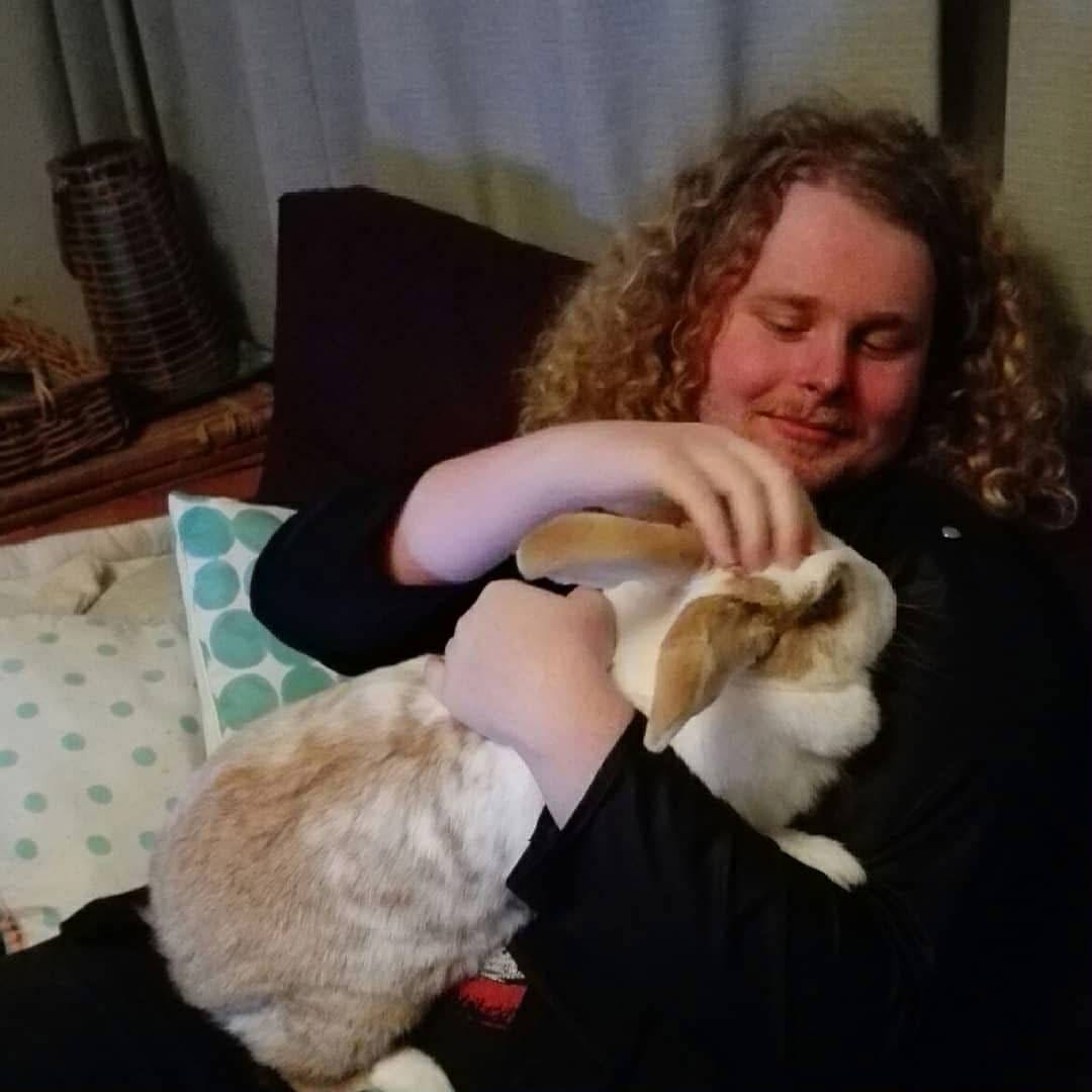 A person with long curly blond hair sits on a couch, smiling softly while holding and gently petting a large white-and-tan rabbit. The rabbit rests comfortably in their arms. A person with long curly blond hair sits on a couch, smiling softly while holding and gently petting a large white-and-tan rabbit. The rabbit rests comfortably in their arms.