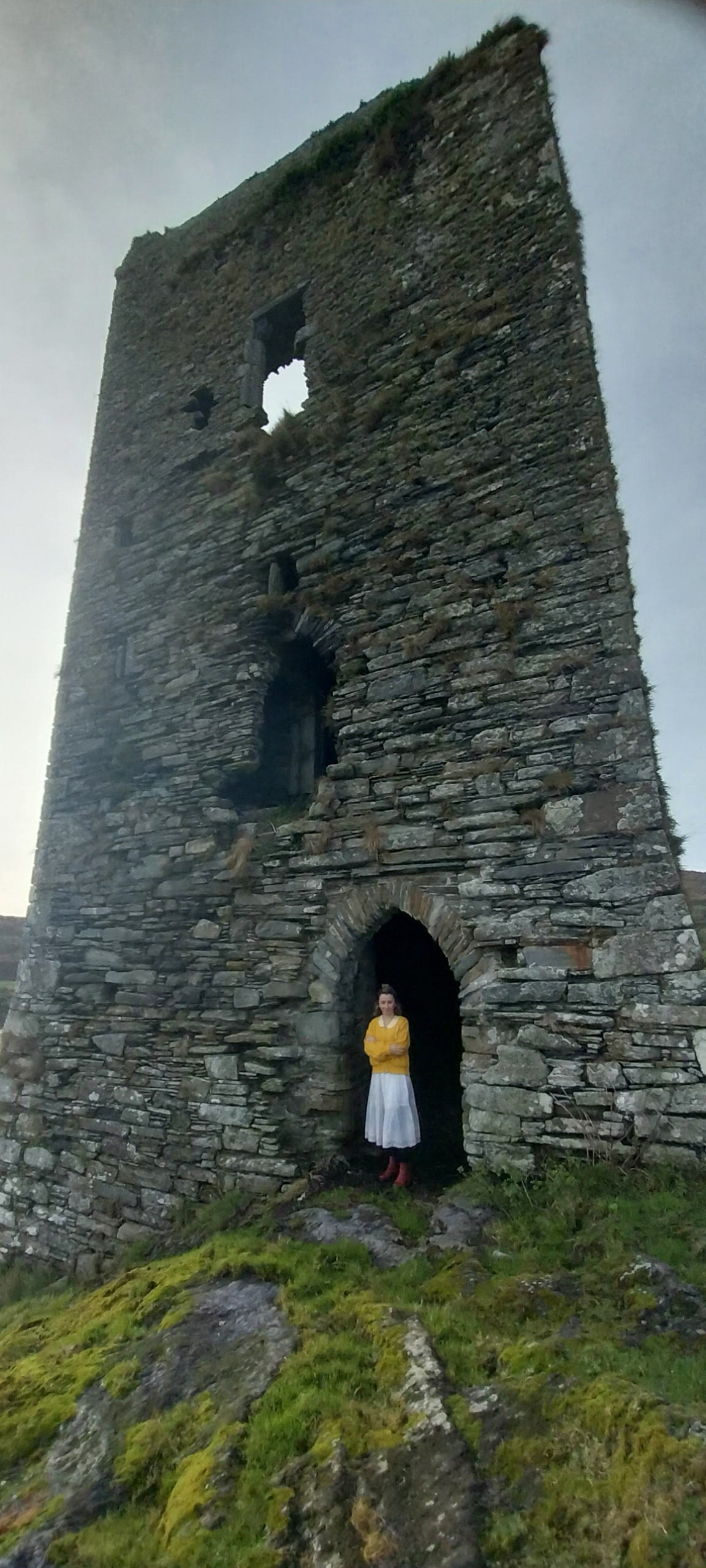 Jennifer in a yellow top and light-colored skirt standing in the arched doorway of an ancient Irish stone tower, with a window opening visible higher up in the weathered stonework.