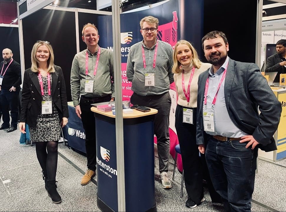 Five Waterstons employees wearing lanyards with QR codes on them stand around a Waterstons branded podium with a large stand in the background.