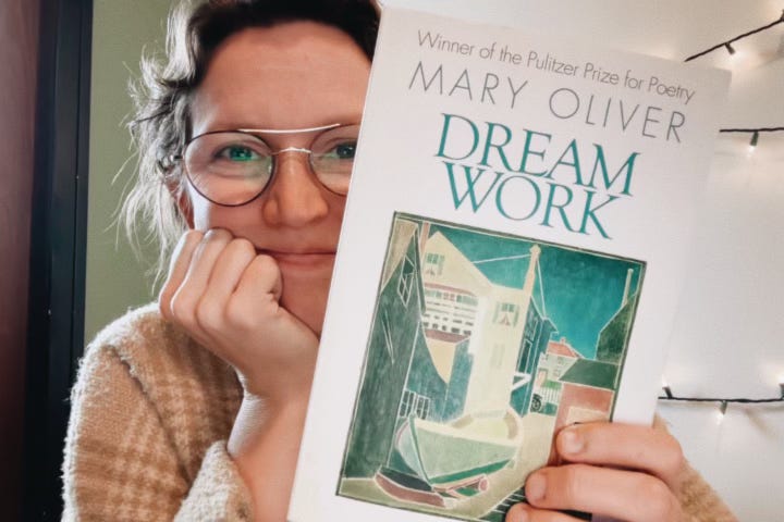 torri in a cream and tan plaid sweater. She is smiling with her head in her hand and holding a copy of Mary Oliver's "Dream Work" to the lens. A small wound on her finger has been blurred out by her photo-editing wife, and this is a fun fact only you who engage with the alt-text will know.