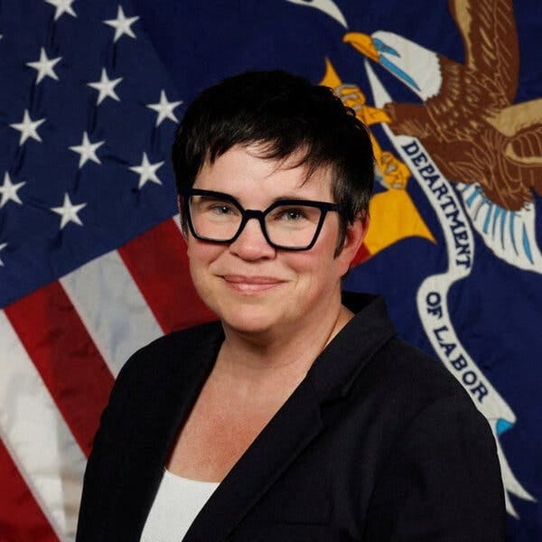 A portrait of Erika McEntarfer, wearing glasses and a dark blazer and sitting in front of a U.S. flag and a flag for the Department of Labor. 